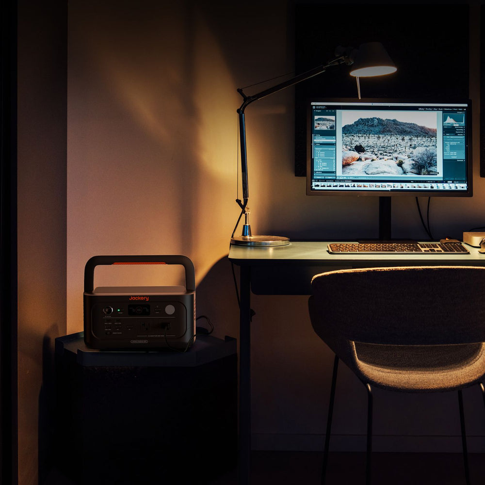 Dark room with a desk setup featuring a computer monitor, keyboard, and lamp with Jackery Explorer 600 v2 in the side