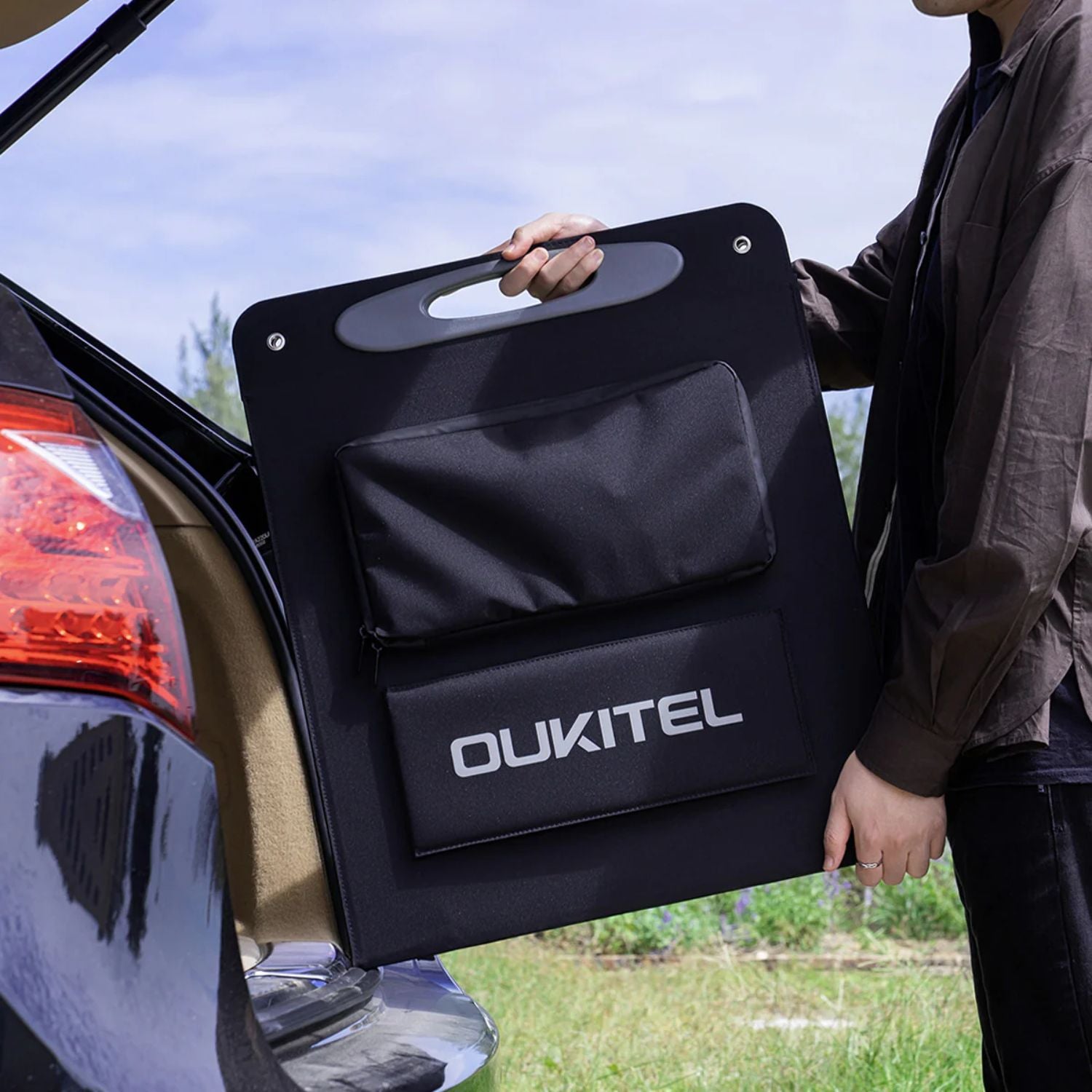 Person holding a OUKITEL 200W Portable Solar Panel against a car exterior.