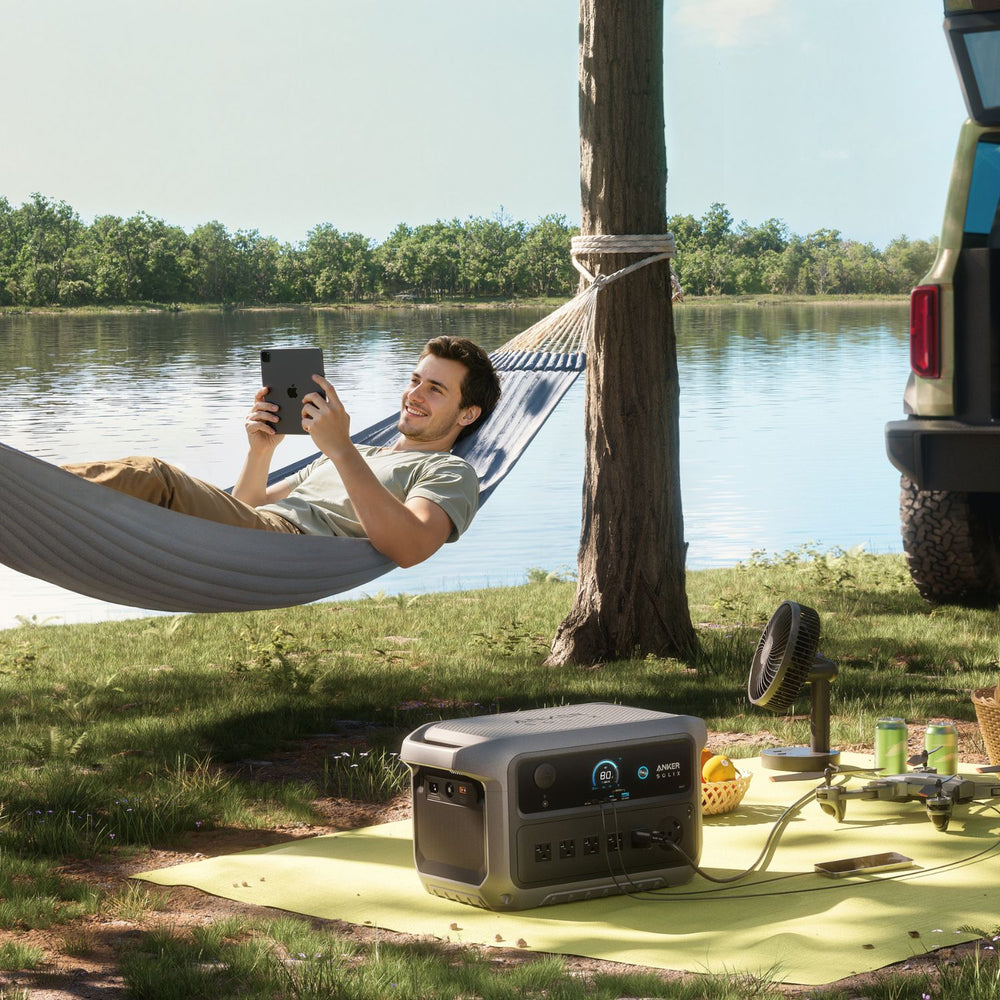 Person in a hammock by a lake using a tablet, with a Anker SOLIX C2000 Gen 2 Portable Power Station on the ground.