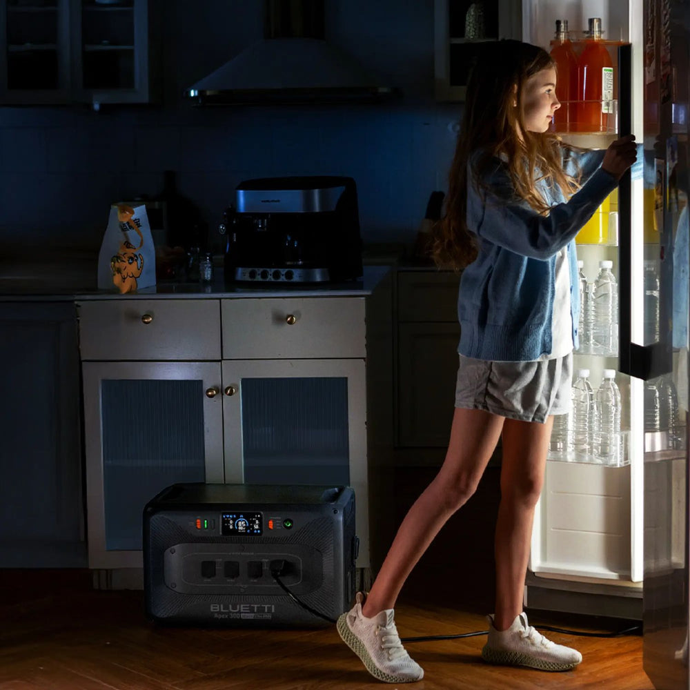 Person standing in a dark kitchen using a BLUETTI Apex 300 Portable Power Station to power a refrigerator.