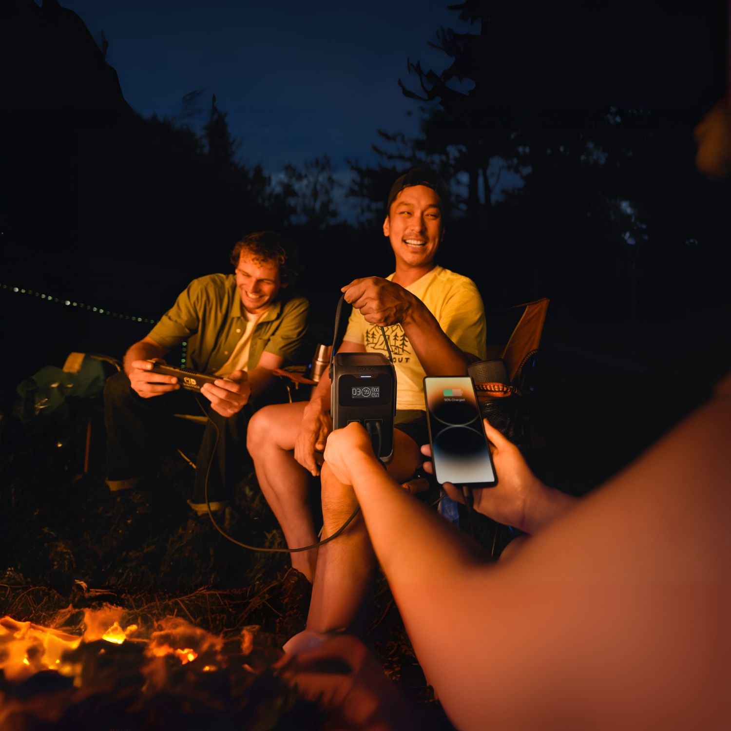 Two people sitting by a campfire at night, one holding an EcoFlow TRAIL 200DC Portable Power Station and the other using a smartphone.
