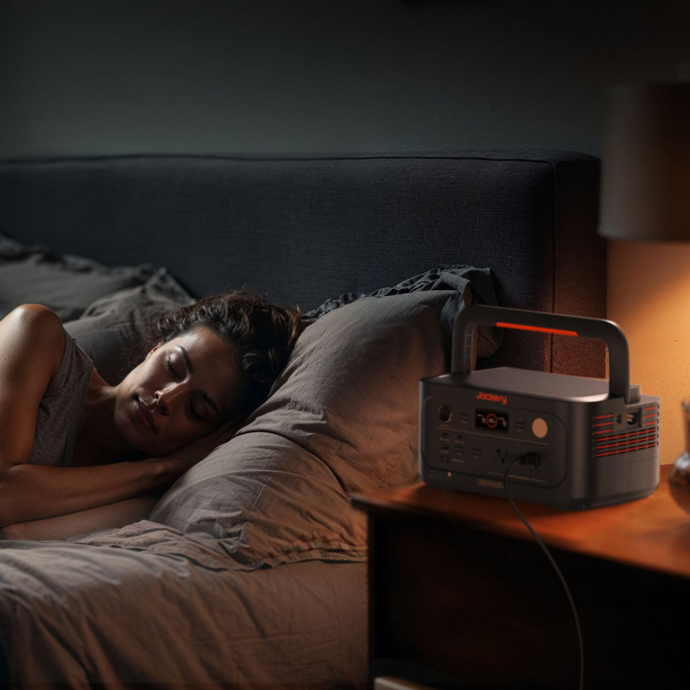 Woman lying in bed with a Jackery Explorer 600 v2 on the nightstand in a dimly lit room.