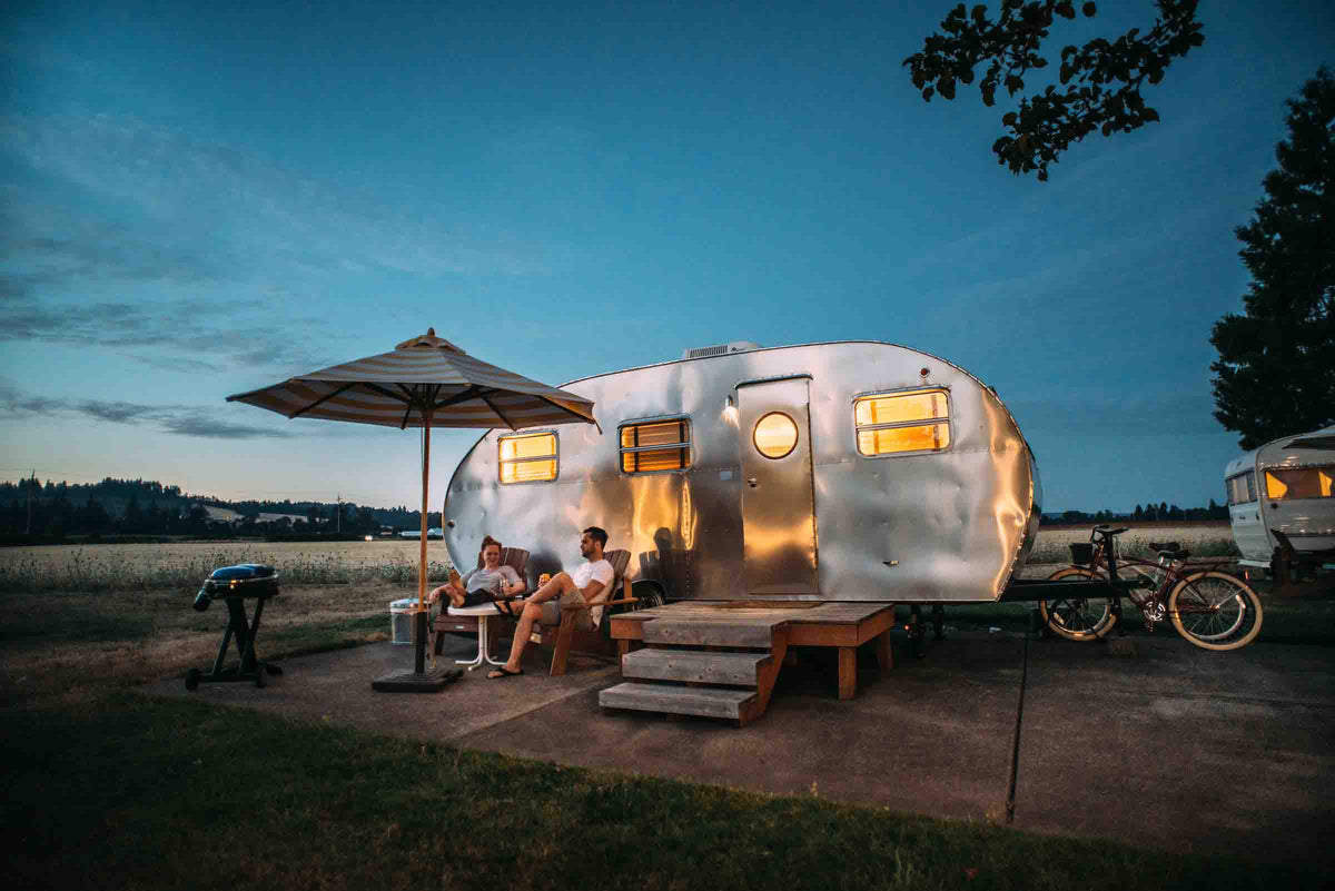 Couple relaxing outside a vintage travel trailer with its lights on at dusk